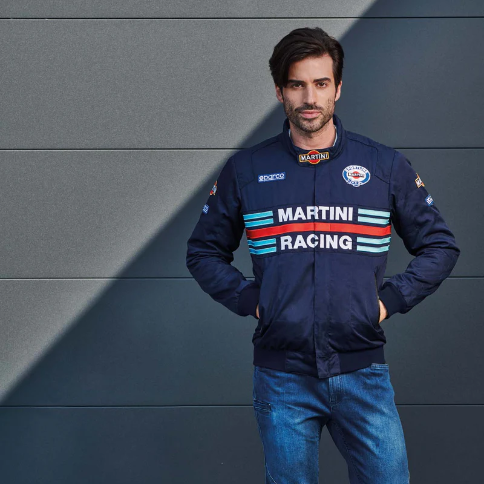 The navy blue Sparco Martini Bomber Jacket from Sparco, featuring Martini Racing and Sparco logos trackside in a lifestyle photograph.
