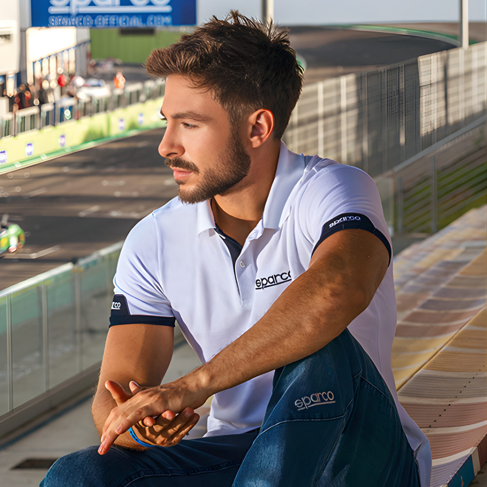 Man sitting on a race track wearing a white polo shirt with 'sparco' branding.