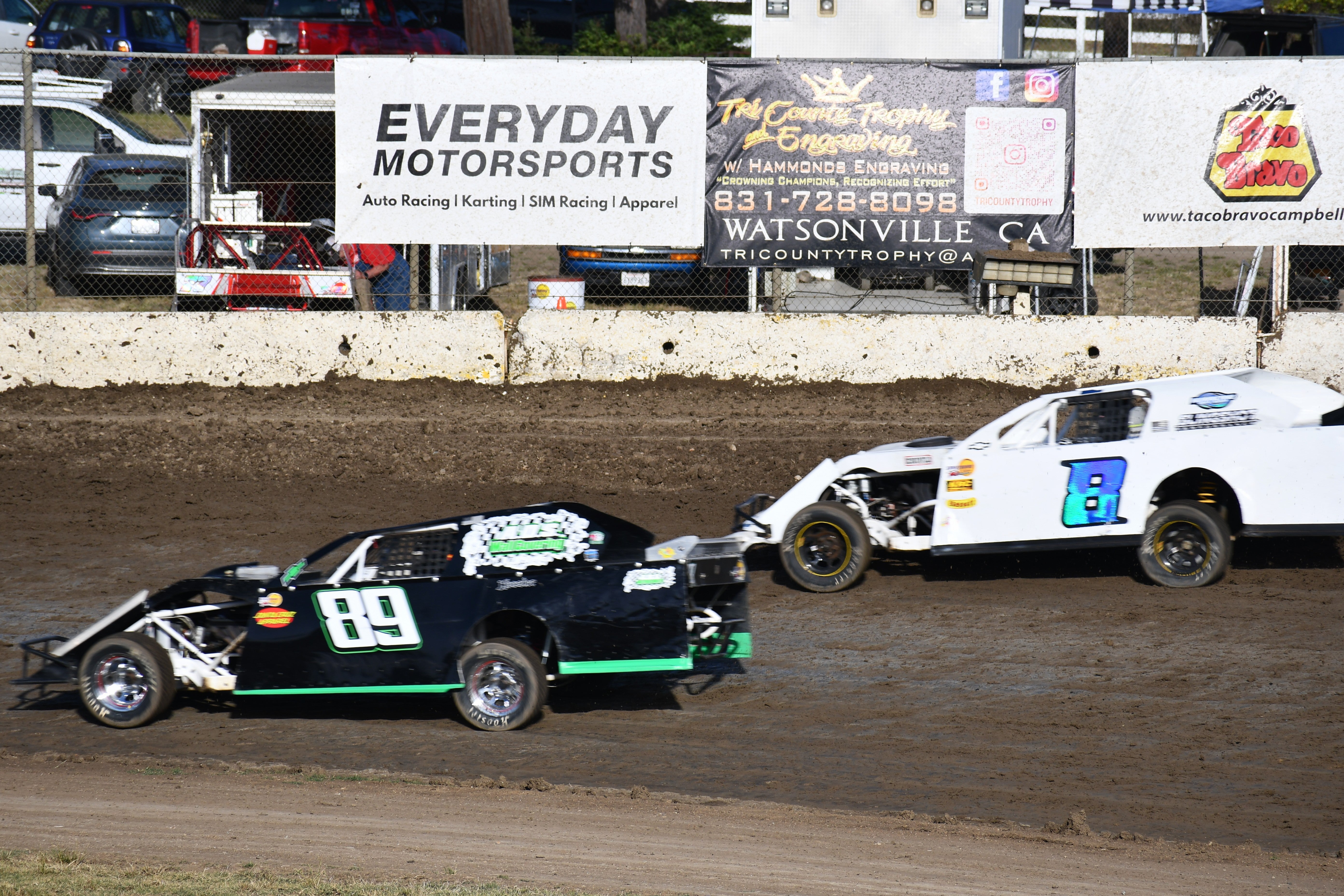 Two race cars on the dirt track at Watsonville Speedway with the Everyday Motorsports Banner in the background.